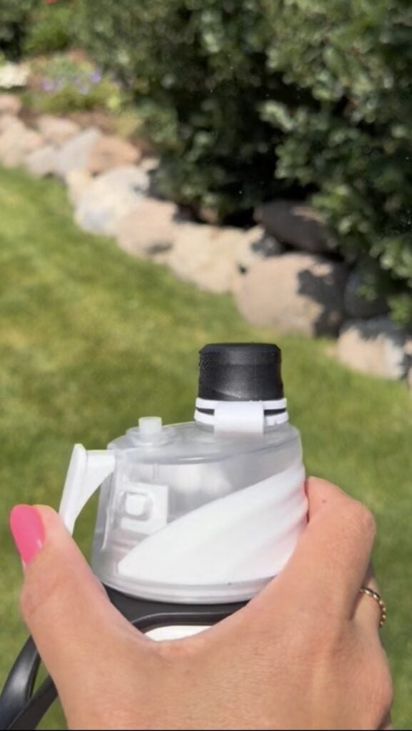 A hand with pink nail polish holds a white and black misting water bottle outdoors, with green grass, rocks, and bushes in the background.