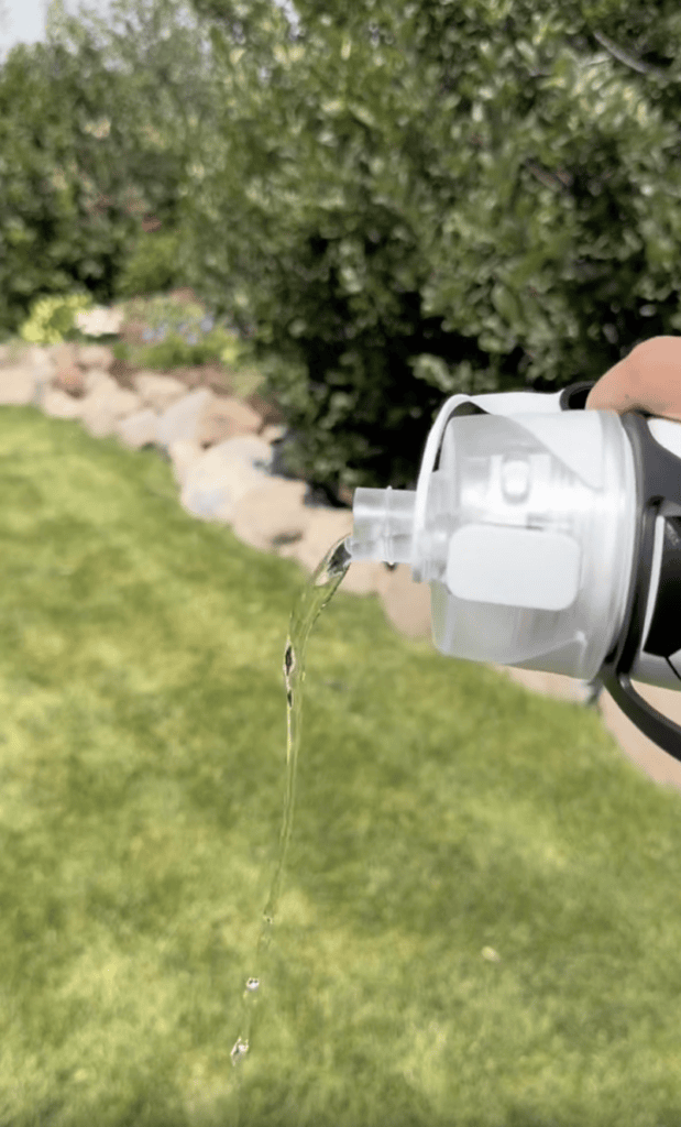 A hand pours water from a clear kids misting bottle outdoors onto green grass, with rocks and trees visible in the background.