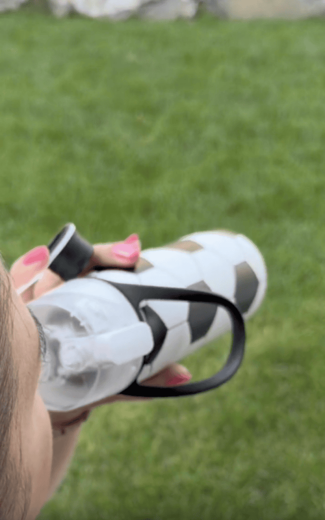 A person with pink nails holds a misting water bottle designed like a soccer ball, about to drink, standing on a grassy area.