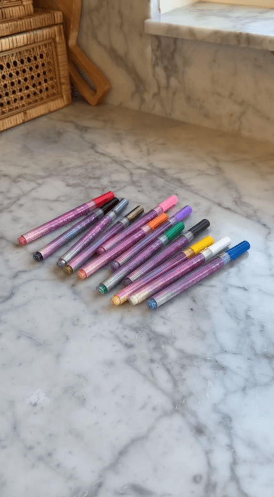 Twelve glittery gel pens in various colors are arranged in two neat rows on a marble countertop, with wicker baskets and a window in the background.