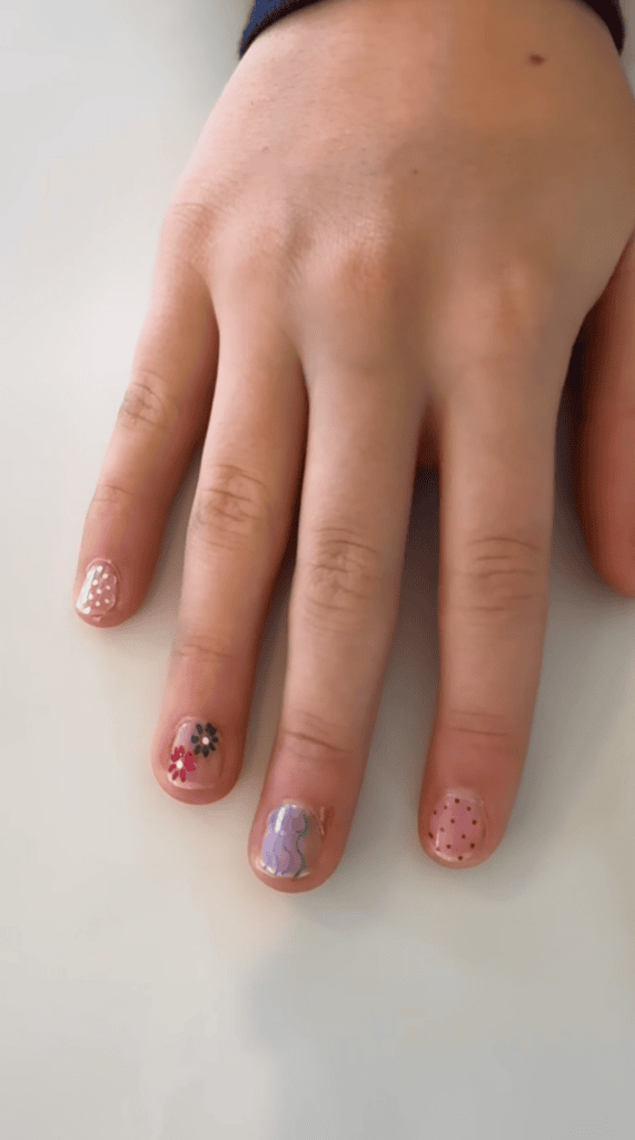A hand with colorful nail art: dots on the pinky and thumb, flowers on the ring finger, wavy blue lines on the middle finger, and red dots on the index finger, all on a pale background.