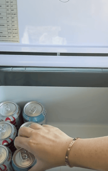 A hand reaches into a refrigerator drawer containing cans of soda, with the fridge temperature guide visible on the inside of the door above.
