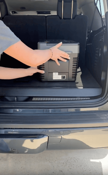 A person places a portable refrigerator or cooler inside the trunk of a black SUV, with the rear seats upright and hatch open.