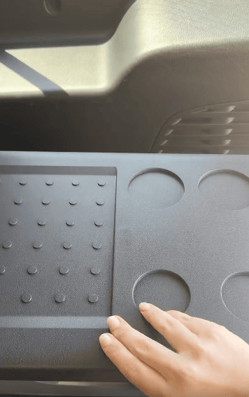A hand touches a textured black dashboard surface inside a vehicle, featuring a dotted pattern on the left and four circular indentations on the right.