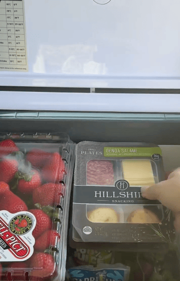 A hand reaches into a fridge drawer, grabbing a Hillshire Snacking plate with salame, cheese, and crackers, next to a container of strawberries.