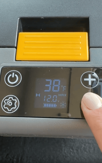 A hand presses a plus button on a digital control panel showing 38°F, 12.0V, and various icons, next to a yellow compartment.