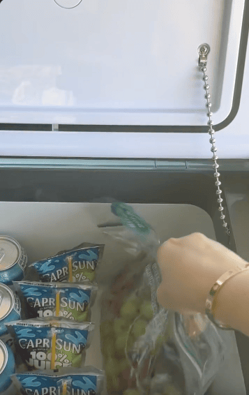 A hand reaches into a cooler containing Capri Sun drink pouches, metal cans, and a bag of green grapes.