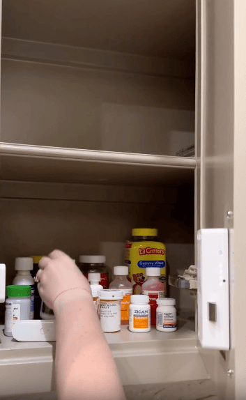 A hand reaches for a bottle among various medicine containers, supplements, and vitamins on a shelf inside a beige medicine cabinet.