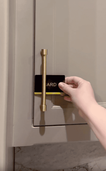 A hand uses a black card to open a cabinet door with a gold handle, suggesting a lock-picking or unlocking action.