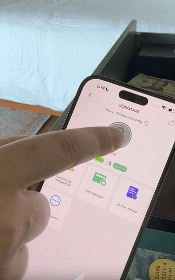 A hand is using a smartphone to control smart home devices, with the screen showing options like unlocking and card management. The background includes a white bed and a nightstand.