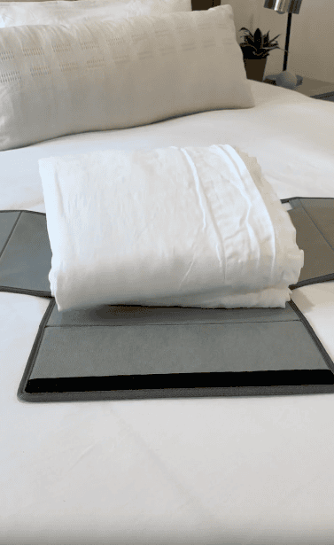 A neatly folded white sheet sits on a grey folding board atop a white bed with white pillows in the background.