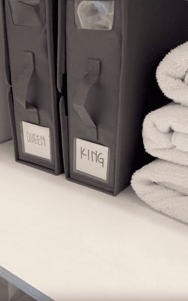 Two labeled grey storage bins—one marked “QUEEN” and one “KING”—sit on a shelf next to neatly folded white towels.