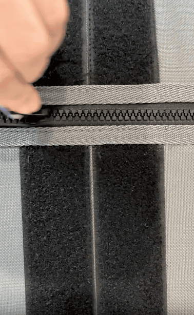 A close-up of a hand pulling a zipper slider along a gray zipper on a black and gray fabric surface.