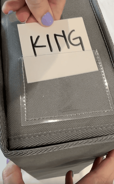 A hand with purple nail polish holds a card labeled “KING” above a clear pocket on a gray fabric storage bin; another hand steadies the bin from below.