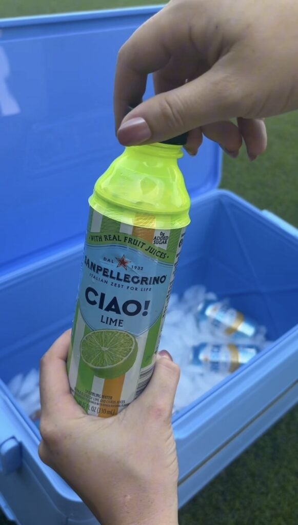 A person opens a can of Sanpellegrino Lime drink over a blue cooler filled with ice and other cans. The scene is outdoors with a grassy background.