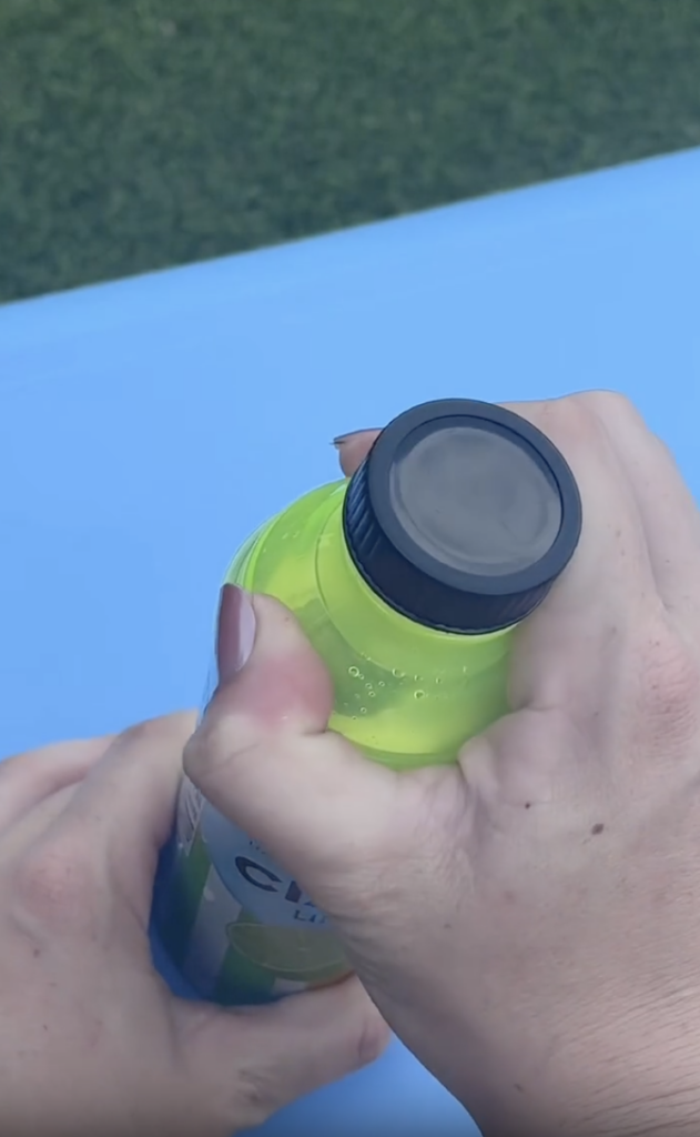 A person is opening a neon green bottle with a black cap, using both hands. The bottle is on a blue surface, with green grass visible in the background.