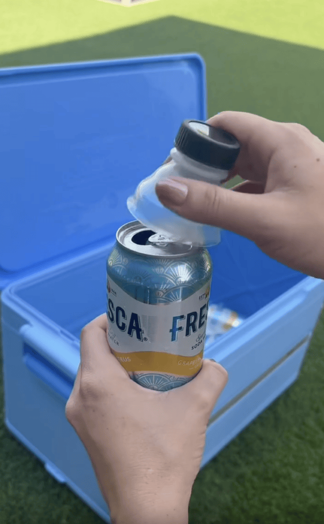 A person sprinkles salt from a shaker onto an open can of Fresca by a blue cooler, with grass in the background.