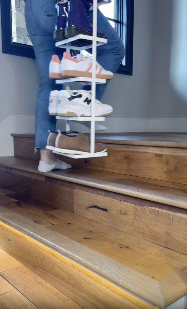 A person wearing jeans and white socks carries a white shoe rack with several pairs of sneakers up wooden stairs, with sunlight streaming onto the steps.