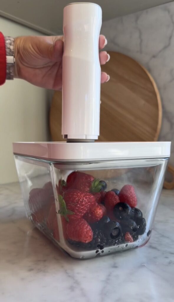 A hand uses a white vacuum sealer on a container filled with strawberries and blueberries, sitting on a marble countertop with a wooden cutting board in the background.