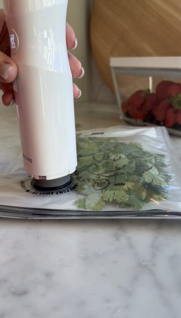 A hand uses a white vacuum sealer on a bag filled with fresh cilantro on a marble countertop. Strawberries in a container are visible in the background.