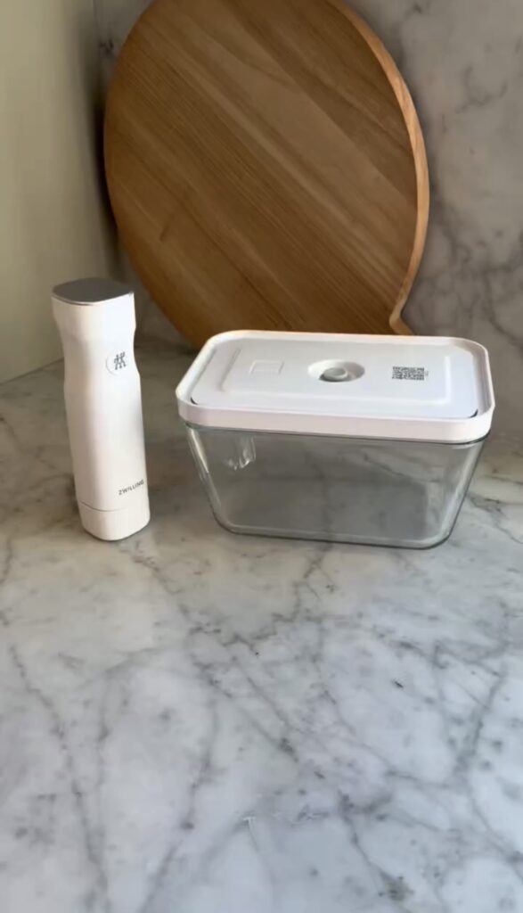 A white handheld vacuum sealer and a rectangular glass container with a white lid sit on a marble countertop, with a round wooden cutting board propped against the wall in the background.