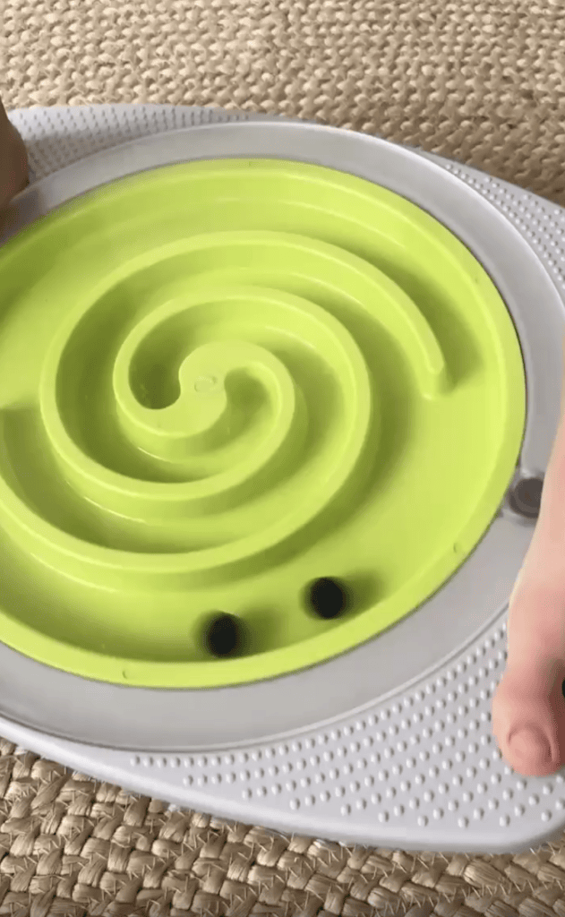 A close-up of hands holding a round maze toy with a green spiral track and two black balls inside, placed on a textured woven surface.